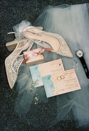 Elegant wedding flat lay with lace heels, floral invitations, pearl earrings, a watch, and tulle veil on gray surface.