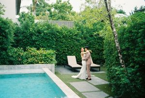 Couple embracing in elegant outdoor garden with turquoise pool, lush green hedges, and stone patio at luxury villa.