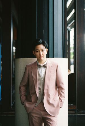 Young man in a pink suit and bow tie leaning against a pillar in an elegant interior setting.