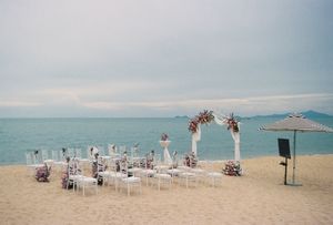 Beach wedding setup with floral arch, white chairs, and ocean backdrop on sandy shore.