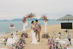 Couple exchanges vows at a romantic beach wedding ceremony with pink floral arch and white chairs by the ocean.