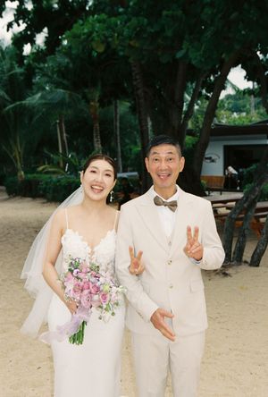 Smiling couple in wedding attire on a tropical beach, bride holding pink bouquet, groom in cream suit making peace signs.