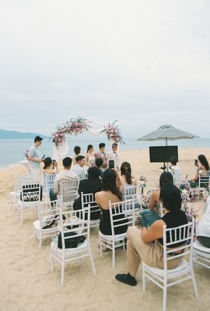 Intimate beach wedding ceremony with white chairs, floral arch, and guests seated by the ocean on a cloudy day.