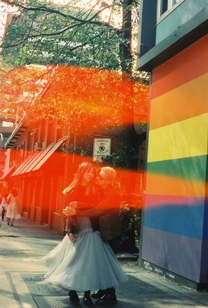 Two people embrace on a city sidewalk beside a rainbow pride flag mural, with warm autumn trees and red light.