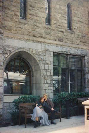 Couple sitting on a wooden bench outside a historic stone building with arched windows and lush greenery.