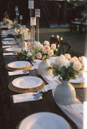 Elegant wedding reception table with gold chargers, white roses, greenery centerpieces, and tall candle holders on a dark wood table.