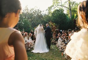 Bride and groom walk down outdoor wedding aisle lined with floral arrangements and guests in warm sunlight.
