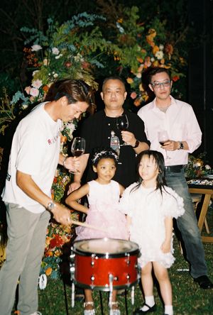 Adults toasting with wine glasses and two young girls in pink dresses stand near a red drum at an outdoor celebration.