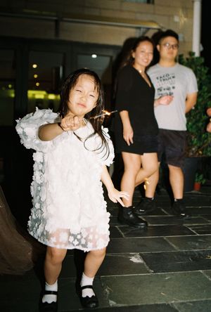 A young Asian girl in a white floral dress holds a sparkler outdoors, with two smiling adults standing behind her.