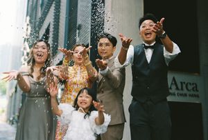 Joyful Asian family celebrating outdoors, tossing confetti in the air near Arca building, dressed in formal attire.