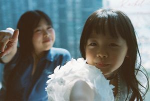Young Asian girl smiling in foreground with woman in blue denim behind her, holding a white feathery object.