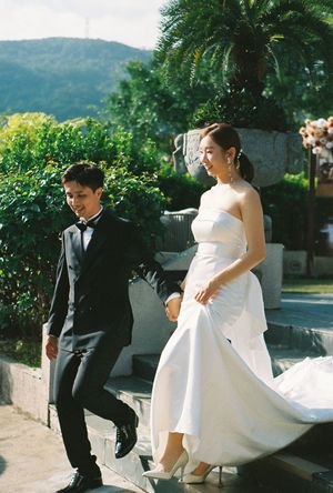 A couple in wedding attire holds hands outdoors, bride in white gown and groom in black tuxedo, with lush greenery behind.