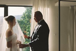 Bride in white gown and veil stands with groom in black suit by a sunlit window with sheer curtains.