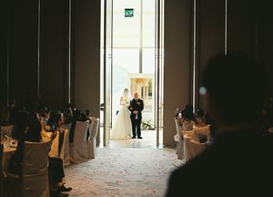 Bride and father walk down the aisle at an elegant indoor wedding ceremony with seated guests.