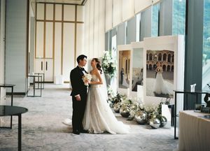 Bride and groom share a kiss in an elegant wedding venue with floral arrangements and framed artwork in the background.