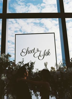 Wedding sign reading Suet & Yik displayed against large windows with blue sky, surrounded by floral arrangements.
