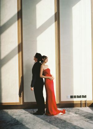 Couple in elegant attire, man in black suit and woman in red gown, standing back-to-back in sunlit hall.