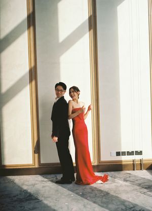 Couple posing elegantly, man in black tuxedo and woman in red strapless gown, standing back-to-back in sunlit hall.