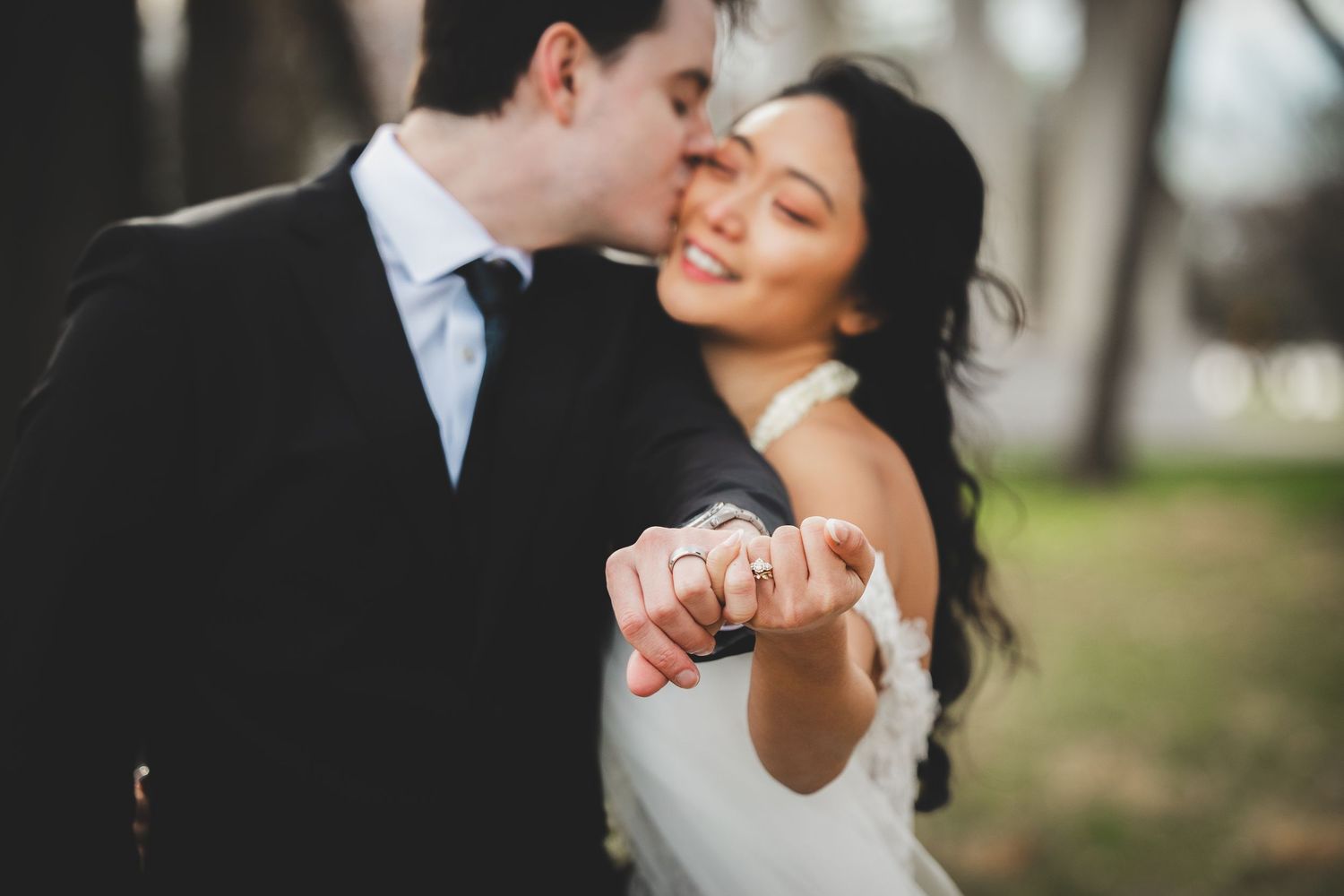 Newlywed couple showing off wedding rings, groom kissing bride on cheek outdoors.