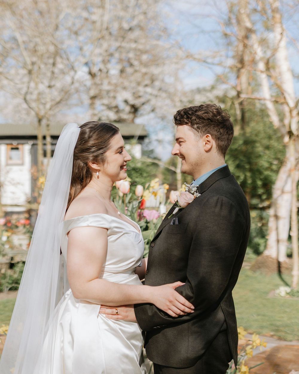 Bride and groom smiling at each other outdoors in a garden wedding setting, bride in white gown with veil.