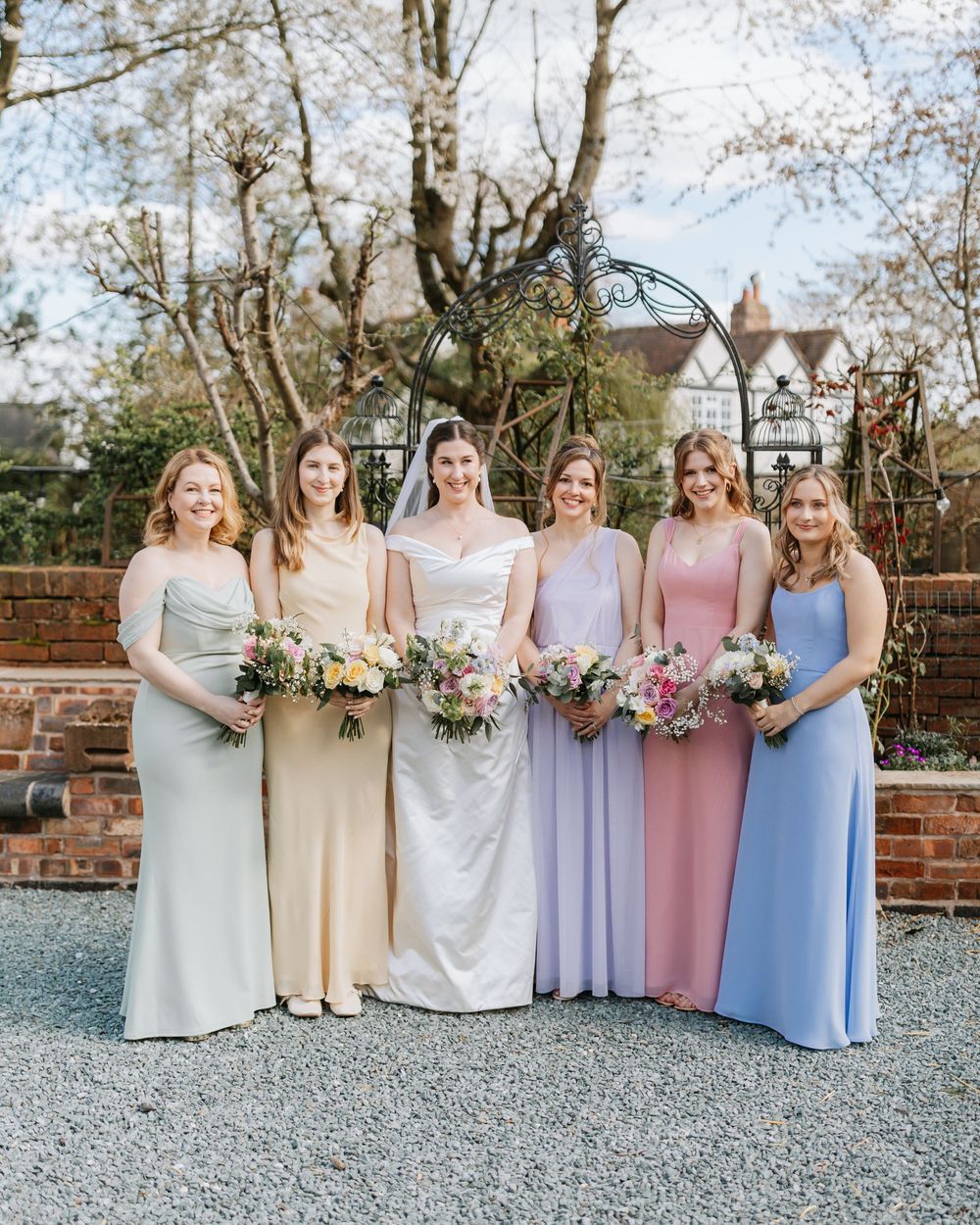 Bride and five bridesmaids in pastel mismatched gowns holding floral bouquets, posing outdoors at a garden wedding venue.
