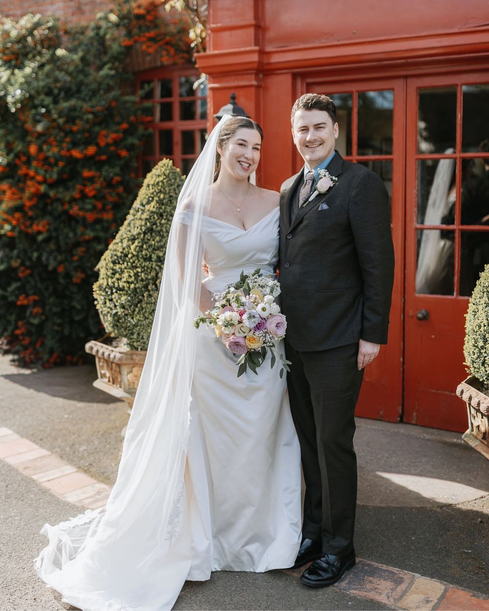 A bride in a white gown with veil holds a floral bouquet beside a groom in a black suit outside a red door.