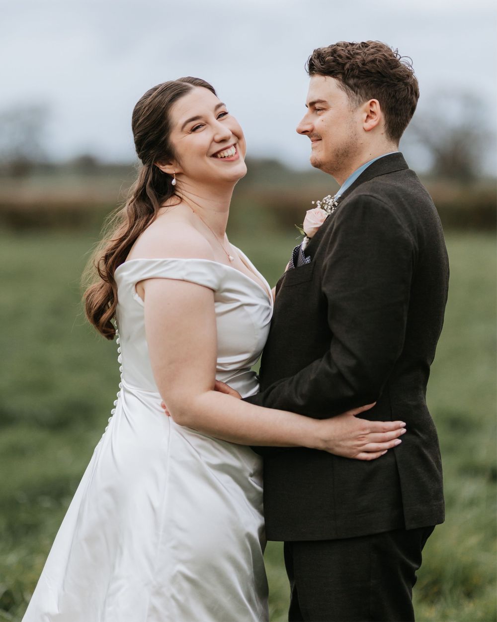 Smiling bride in off-shoulder white gown embraces groom in black suit in green field.