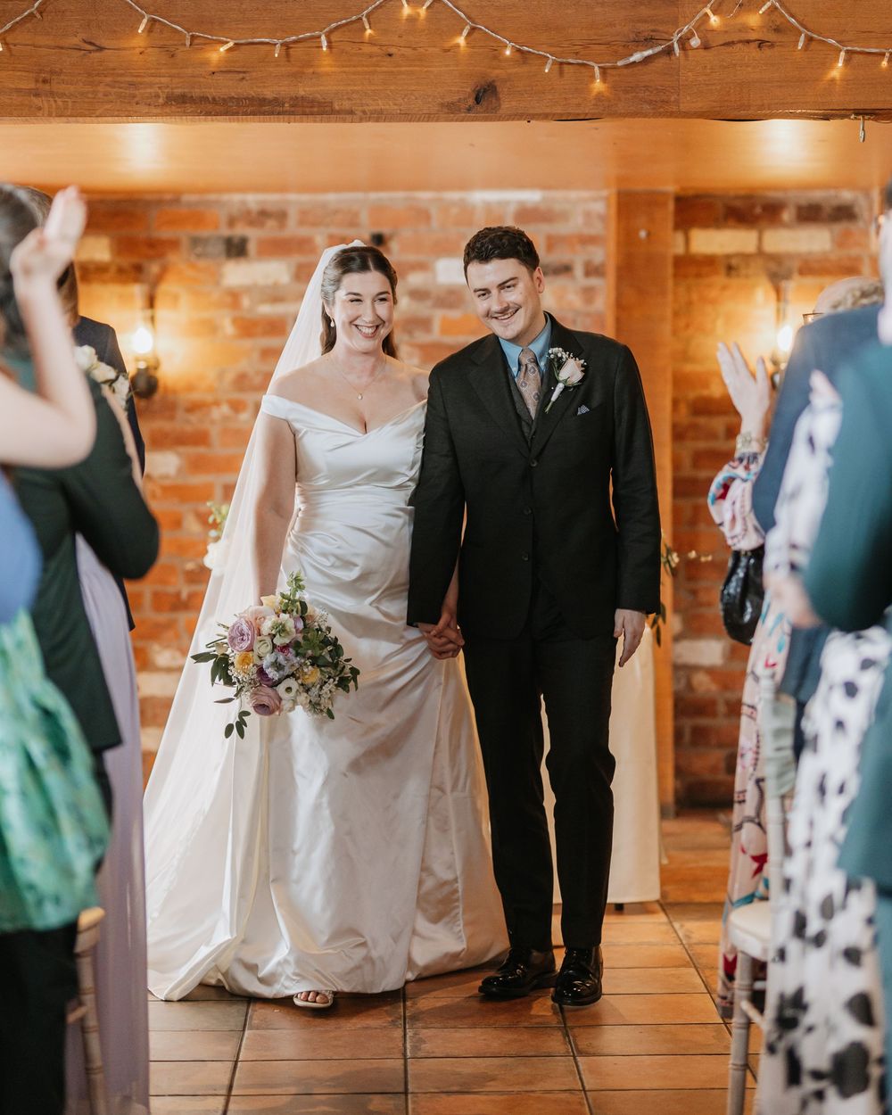Smiling bride and groom walk down aisle in rustic brick venue with fairy lights after wedding ceremony.