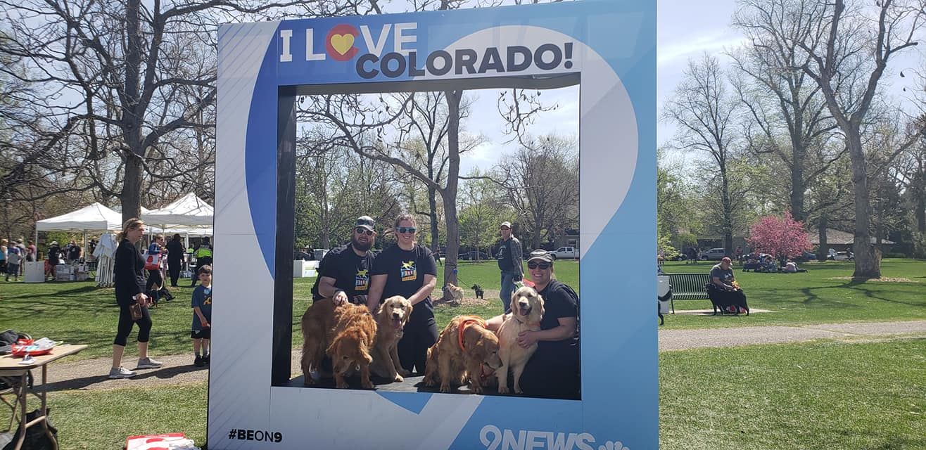 Two people pose with golden retrievers in an I Love Colorado photo frame at an outdoor park event on a sunny spring day.