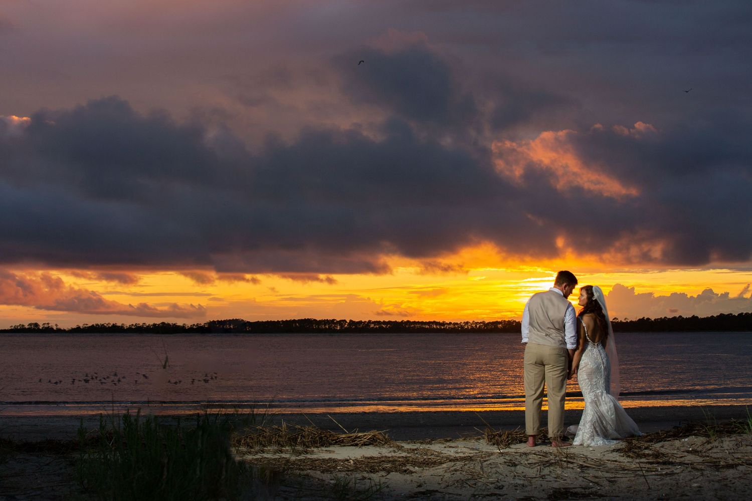Couple in wedding attire silhouetted against a vibrant sunset over calm water on a sandy beach.