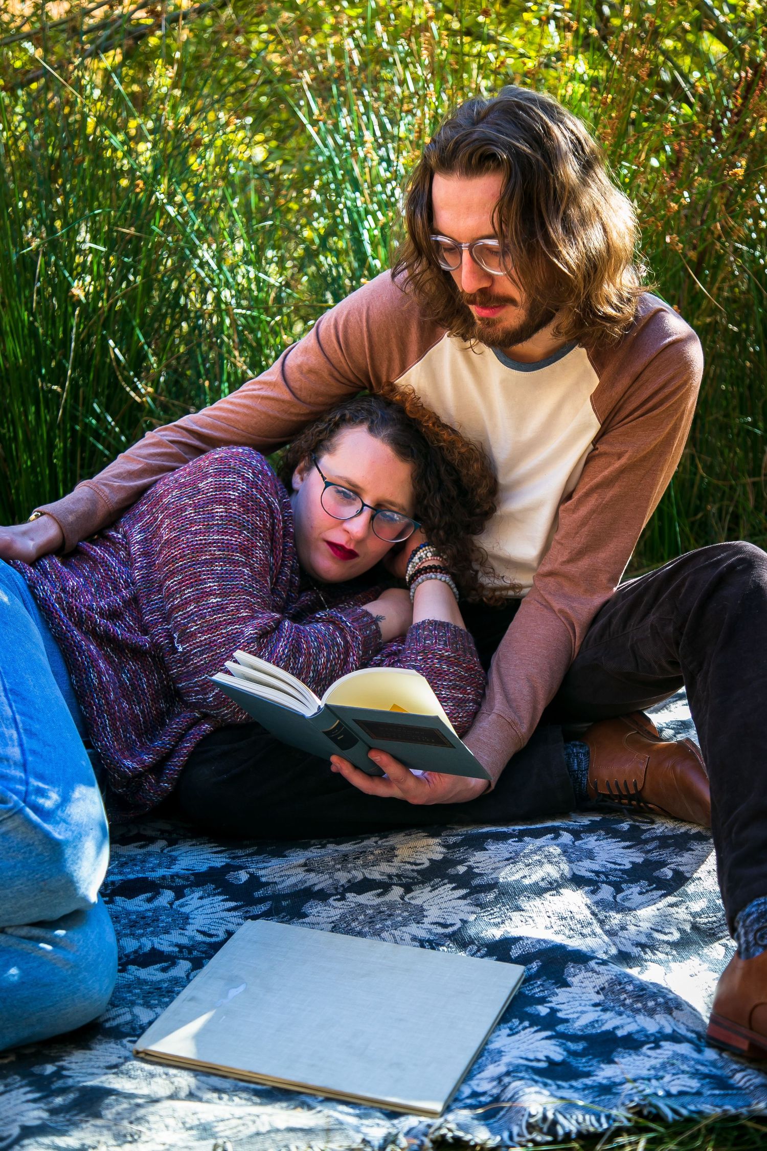 Couple reading a book together outdoors on a blanket surrounded by lush green plants on a sunny day.