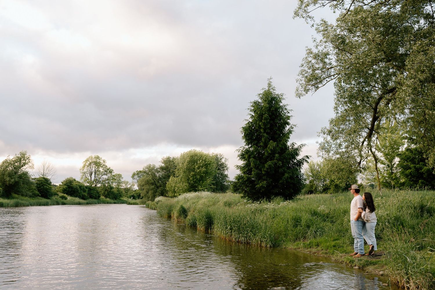 Couple standing by a peaceful river surrounded by lush green trees and reeds under a cloudy sky.