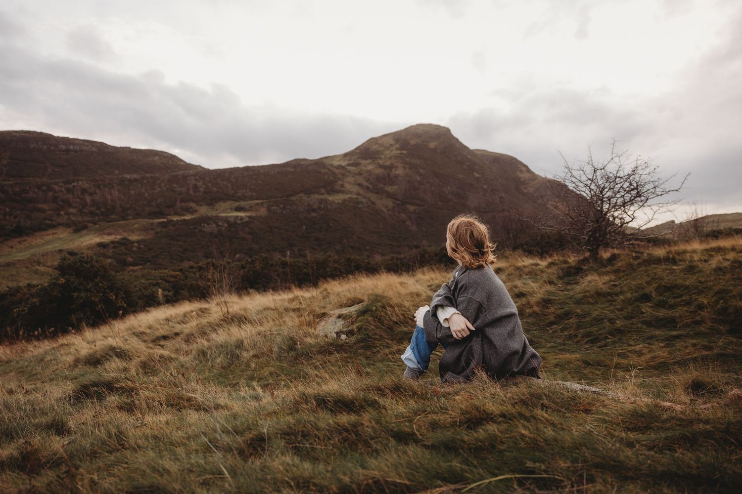 Person sitting on a grassy hillside gazing at rugged brown mountains under a cloudy sky in a remote landscape.