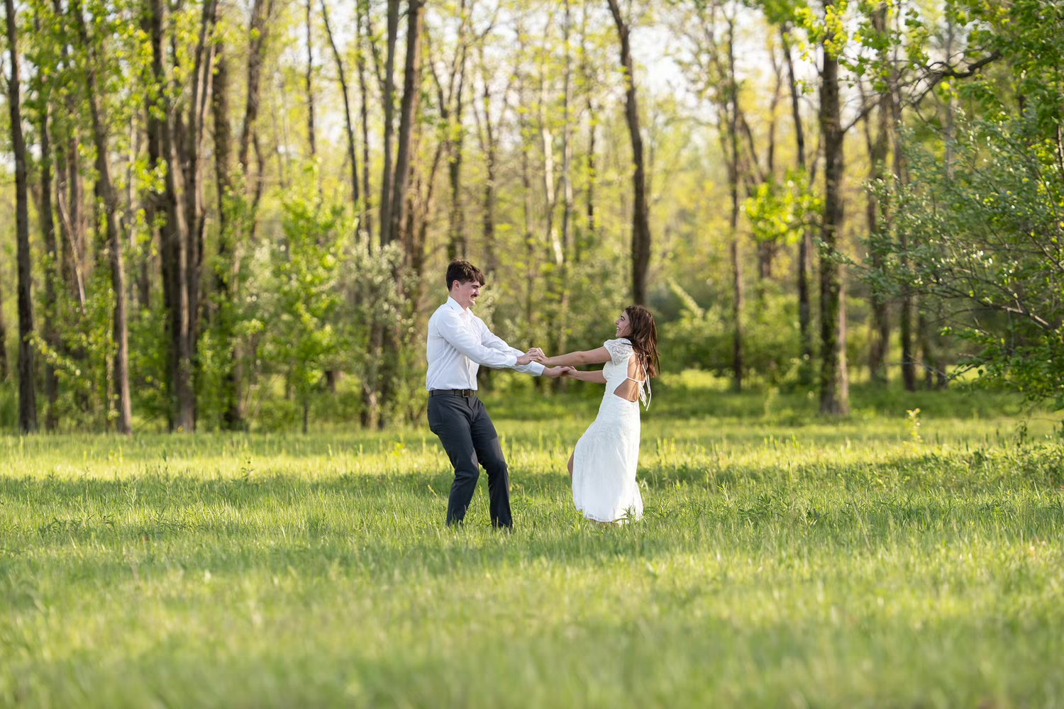 Couple dancing joyfully in a sunlit green meadow surrounded by lush trees, at Alum Creek Beach for Engagement Photos