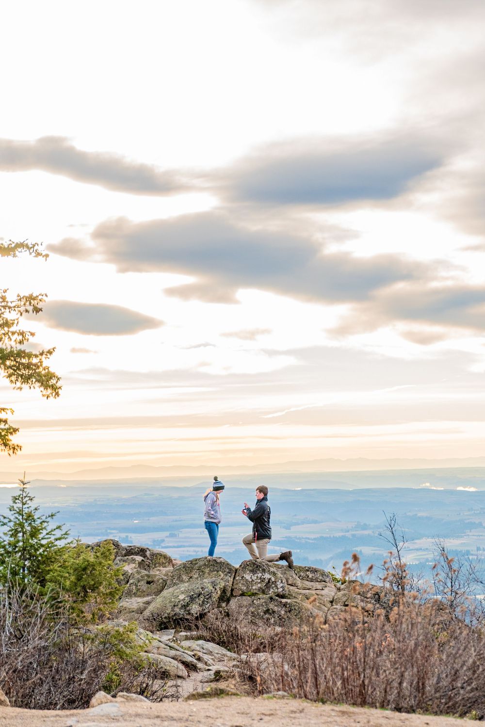 Two hikers stand on a rocky mountain summit overlooking a vast valley landscape at sunset with dramatic cloudy skies.