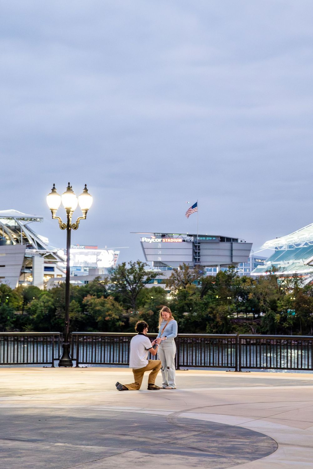 Man proposes on one knee to woman on outdoor plaza at dusk with stadium and American flag in background.