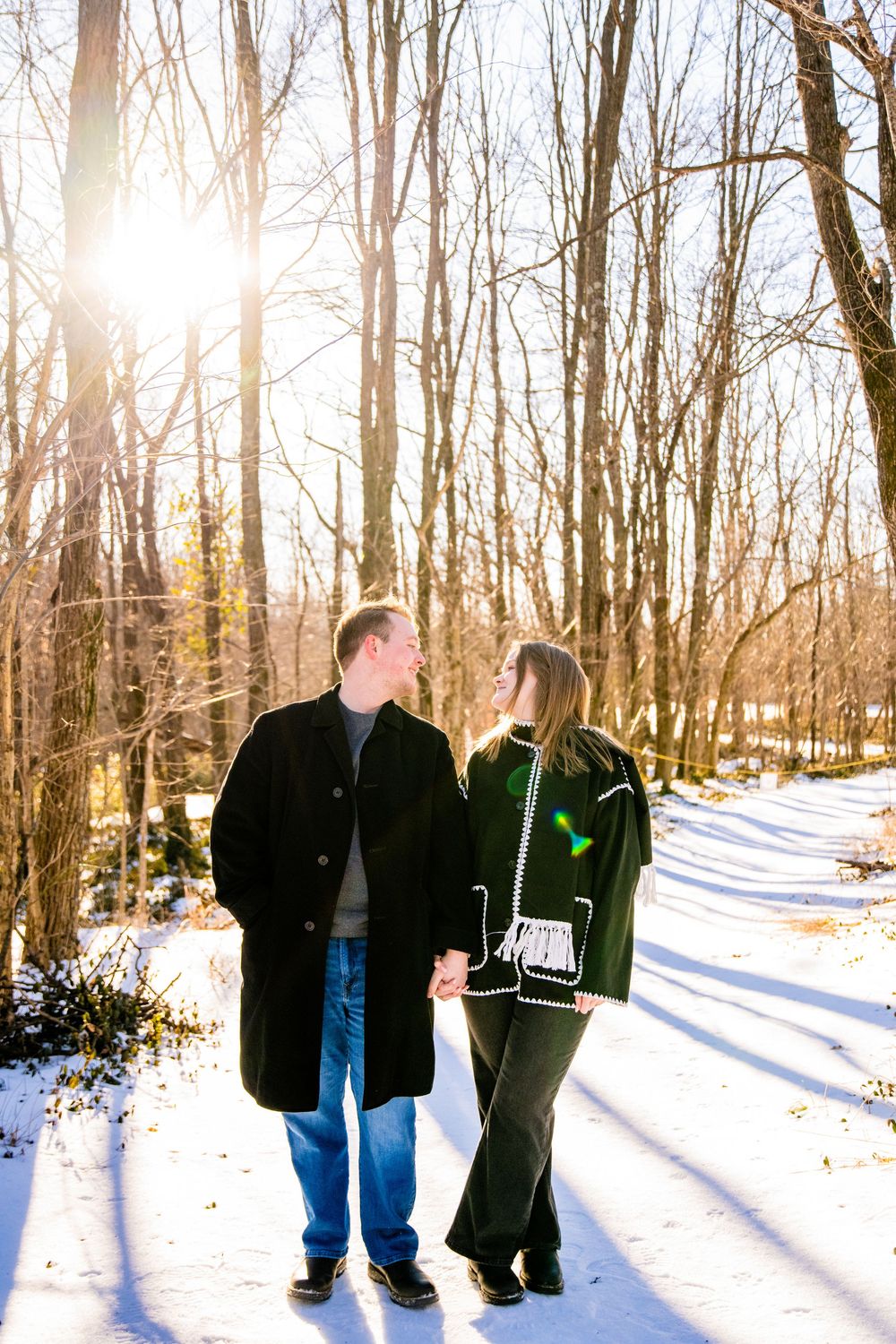 Couple holding hands on a snowy forest path in winter, backlit by golden sunlight through bare trees.