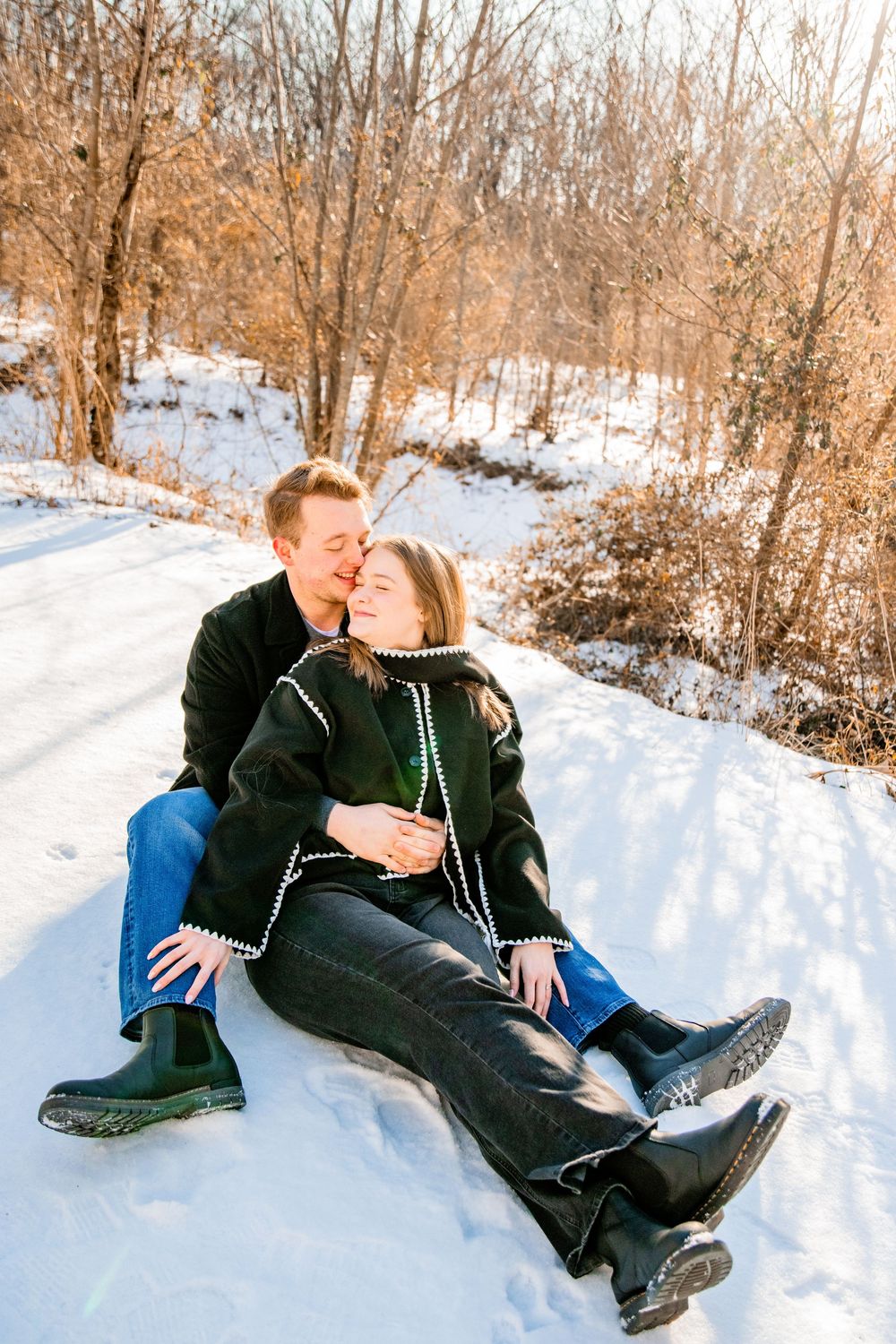 Couple sitting together on snowy ground in winter, wearing black coats and jeans, sharing a romantic moment outdoors.