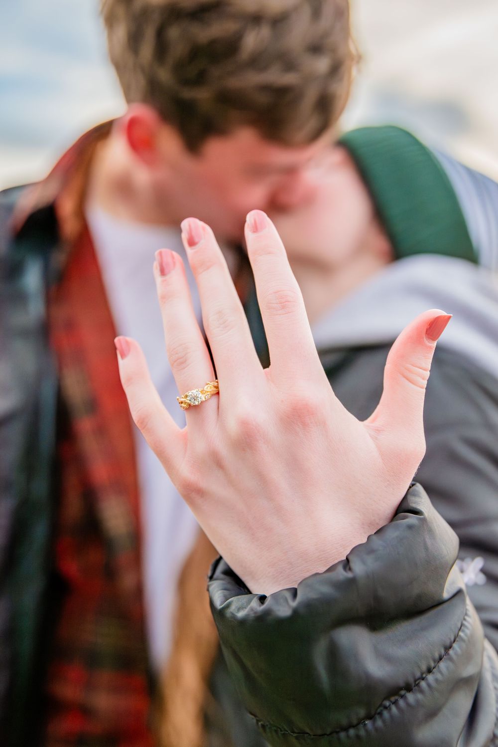 Couple kissing while showing off a gold engagement ring with red manicured nails in the foreground.