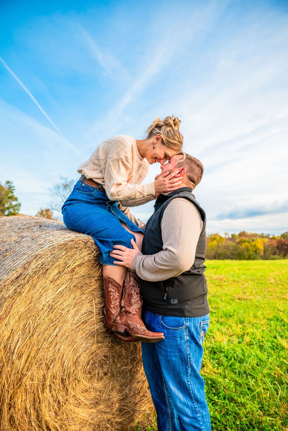 Couple sharing a romantic moment leaning against a hay bale in a green field under a blue sky.