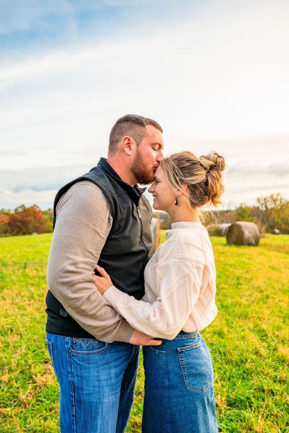 A couple shares a romantic kiss in a sunlit green field with hay bales, the woman in a white sweater and jeans.