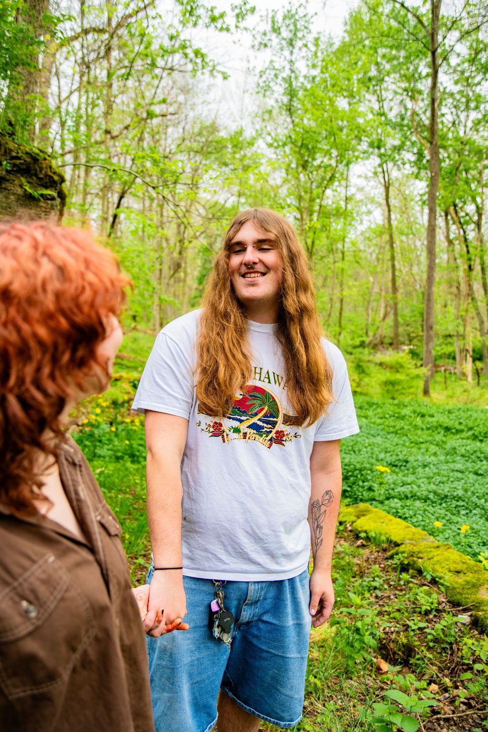 Young man with long auburn hair smiles warmly wearing a graphic tee in a vibrant green woodland.