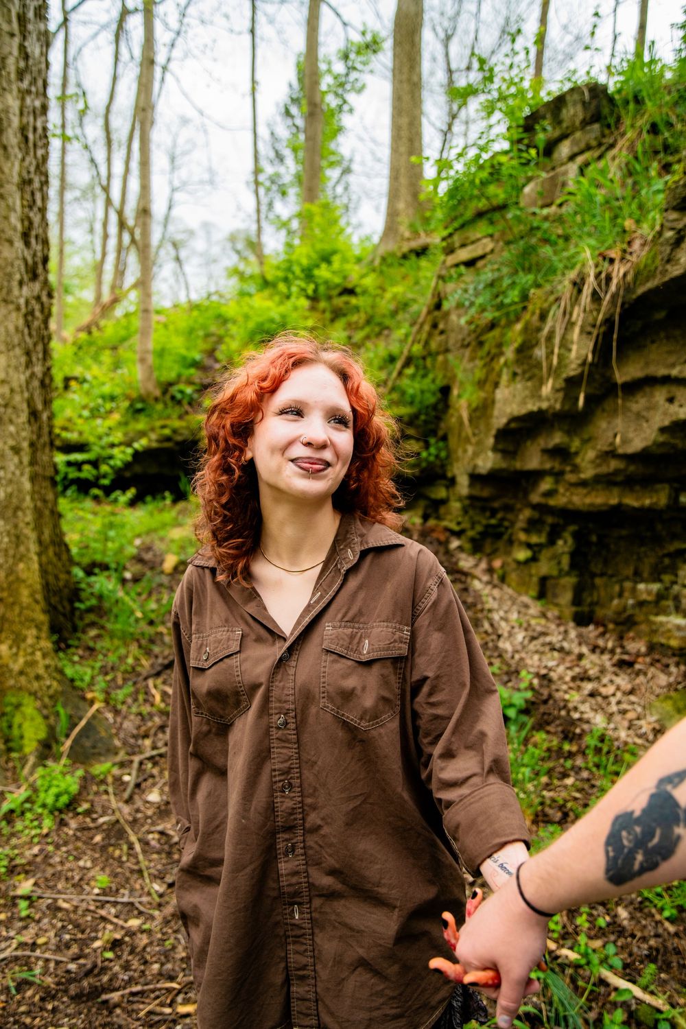 A red-haired woman in a brown shirt smiles while holding hands in a lush forest with rocky cliffs.