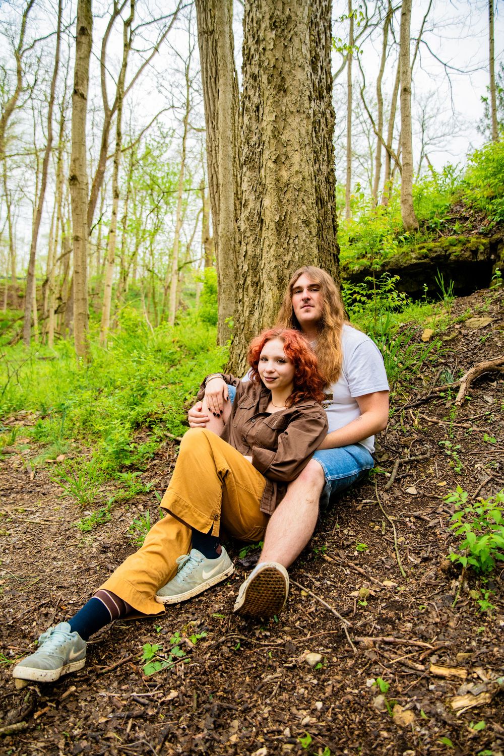 A couple sits together against a tree trunk in a green woodland setting, sharing a tender moment.