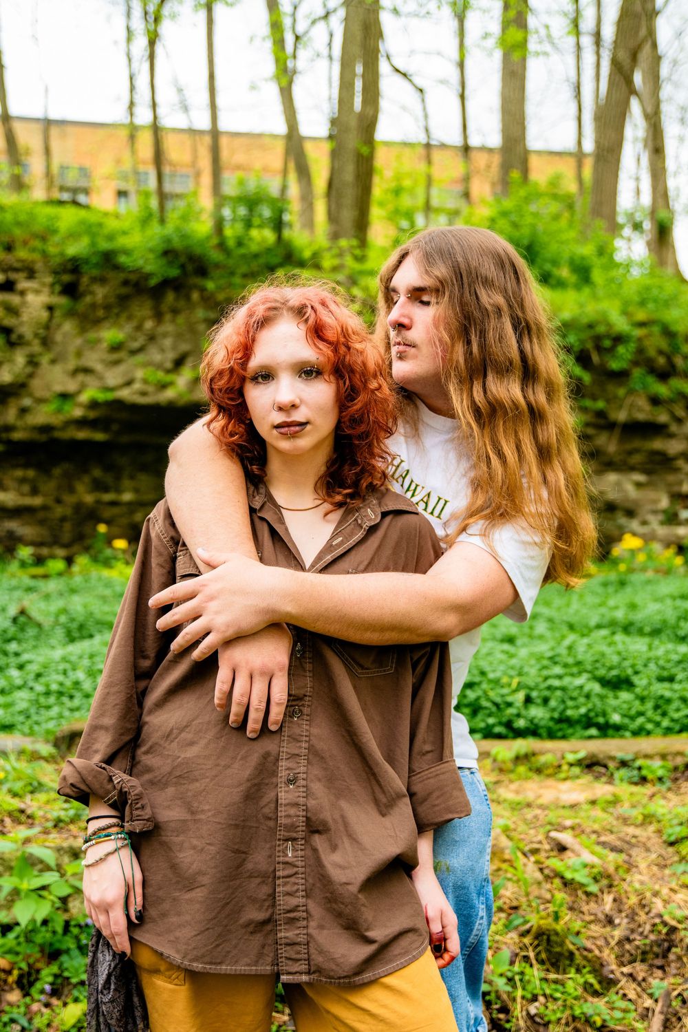 Young couple posing outdoors in a green forest, man embracing woman with red curly hair.