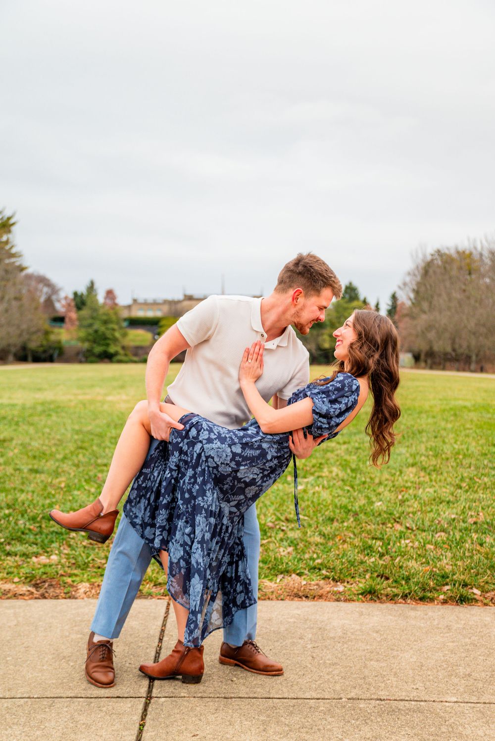 A couple shares a romantic dip pose outdoors, with the man in a white polo lifting the woman in a blue floral dress.