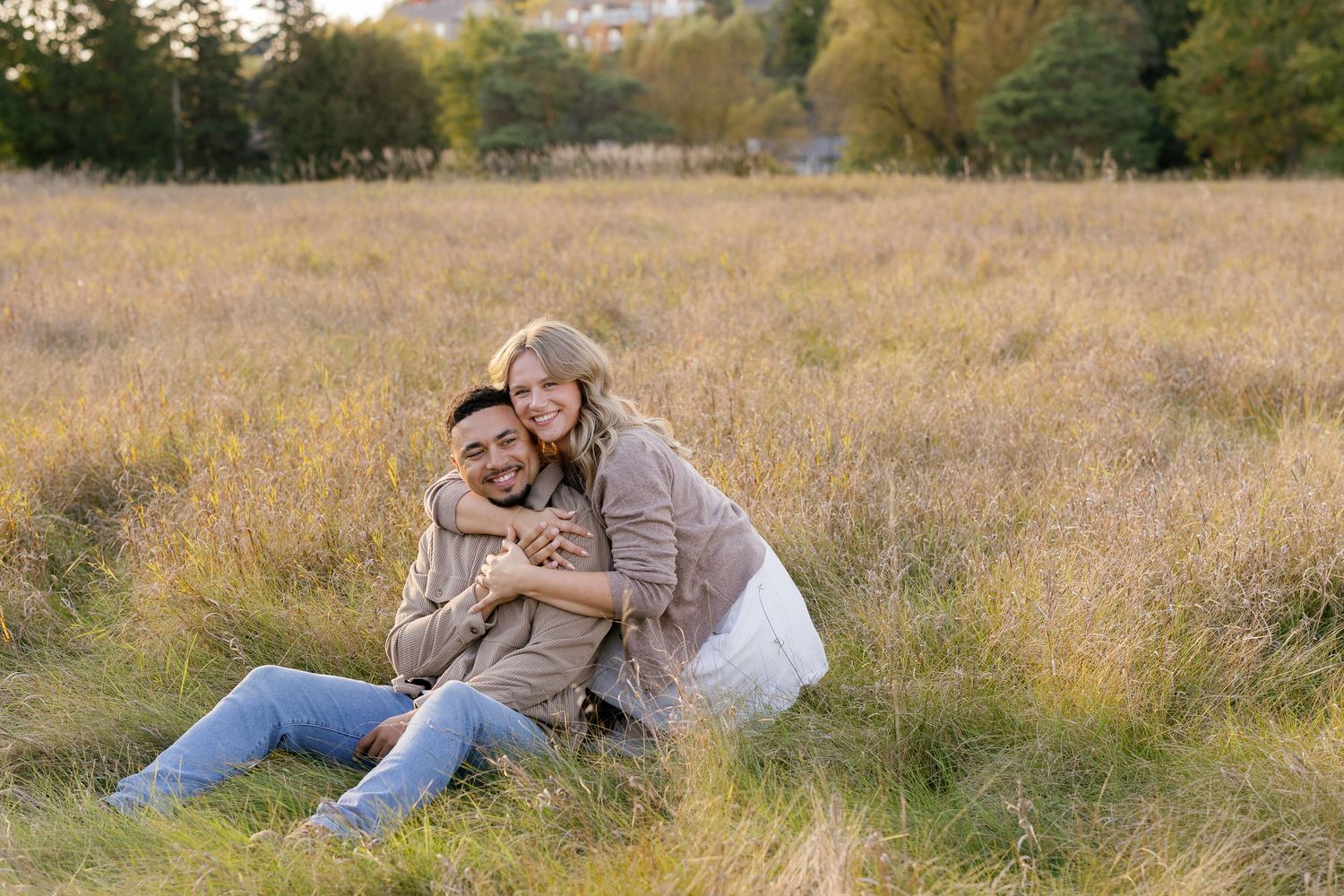 Couple sitting in golden grass field, woman hugging man from behind, smiling outdoors in autumn park.