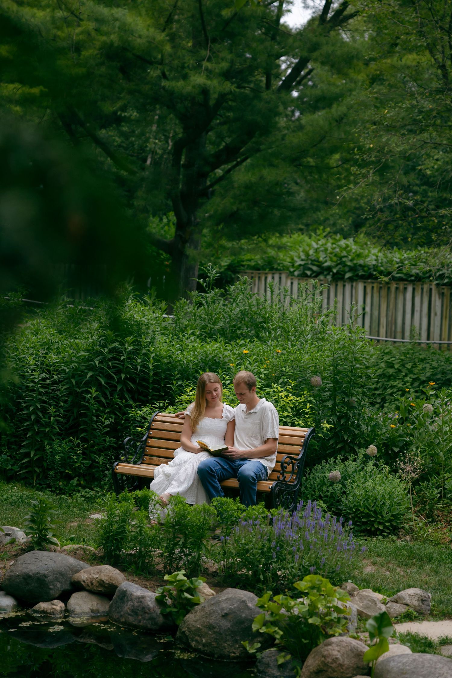 A couple sits together on a wooden bench surrounded by lush green garden plants near a rocky pond.