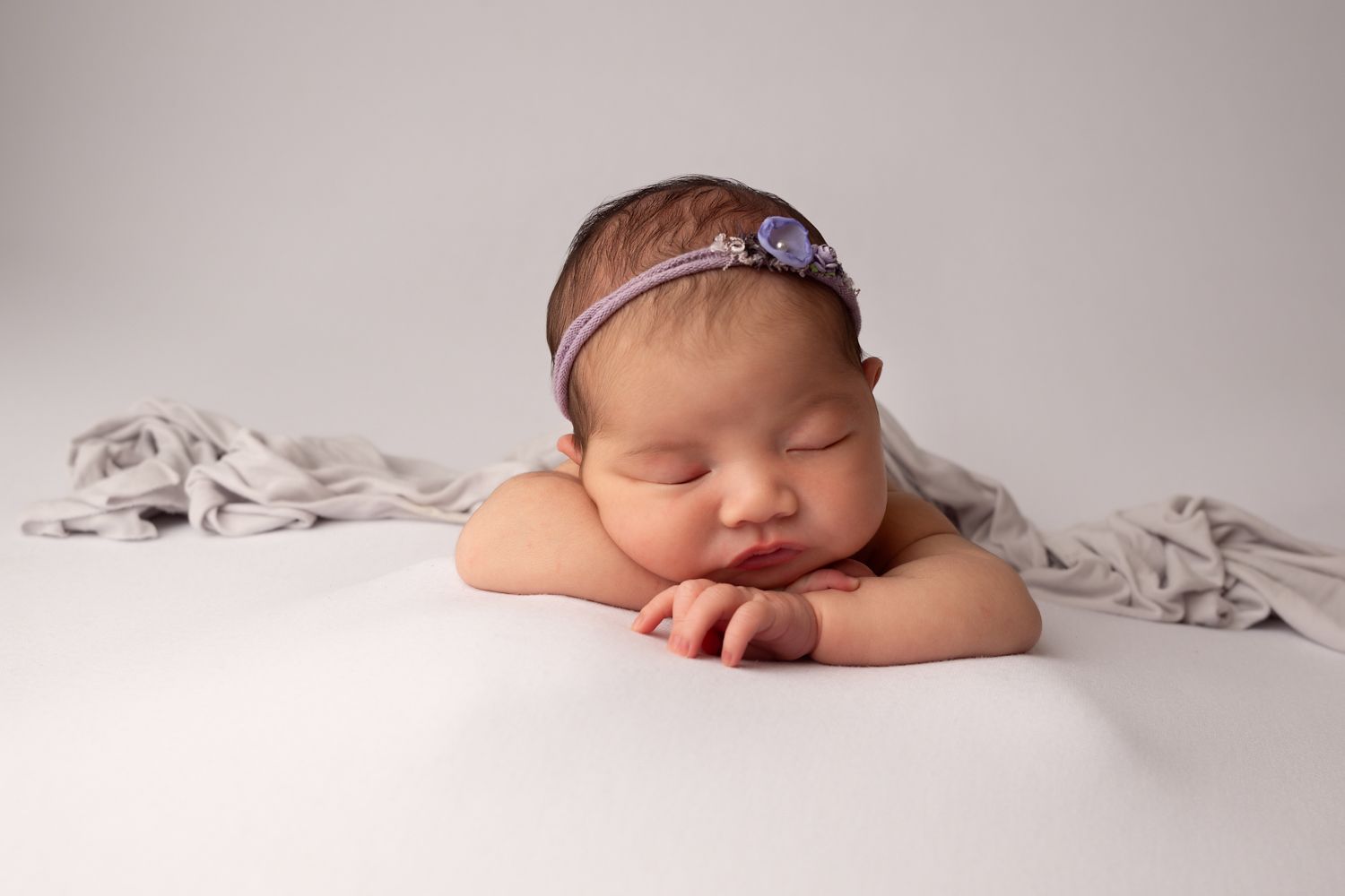 Newborn baby girl sleeping peacefully with chin resting on hands, wearing a purple floral headband on white backdrop.