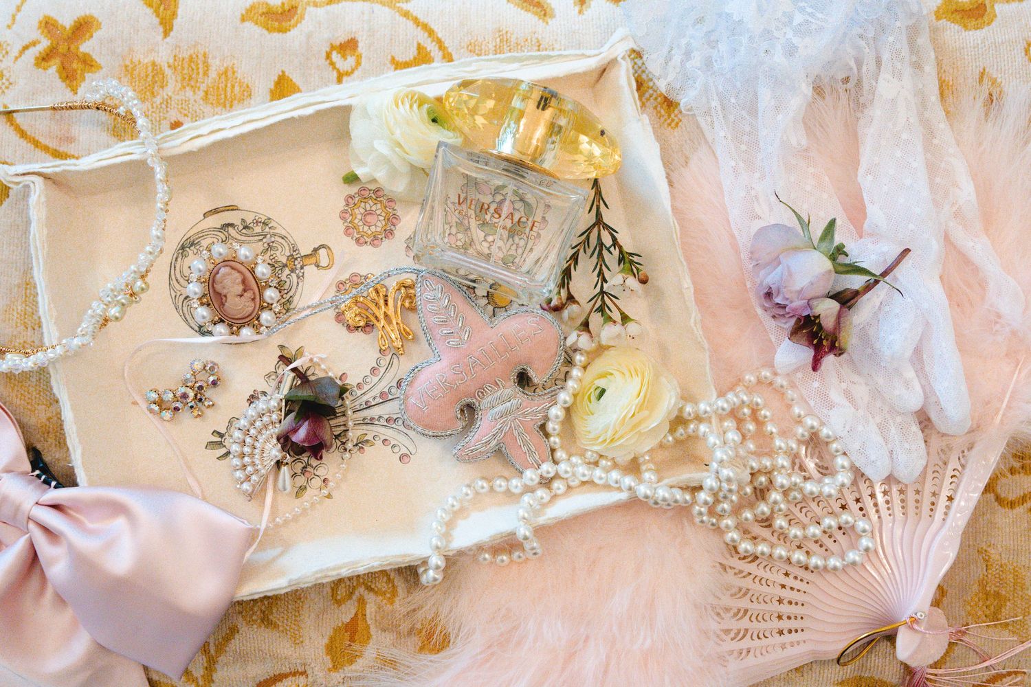 Elegant flat lay of pearl jewelry, vintage brooches, perfume bottle, and white flowers on a pink silk tray.